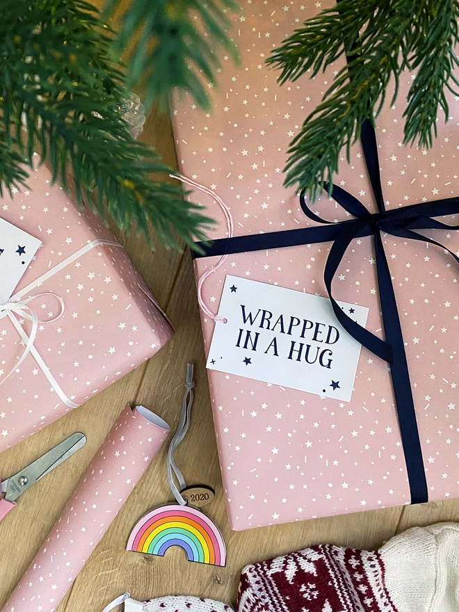 A gift wrapped in a gentle pink start design wrapping paper, tied with navy blue ribbon, with a gift tag that reads "Wrapped in a hug" is on a wooden floor beneath a Christmas Tree.