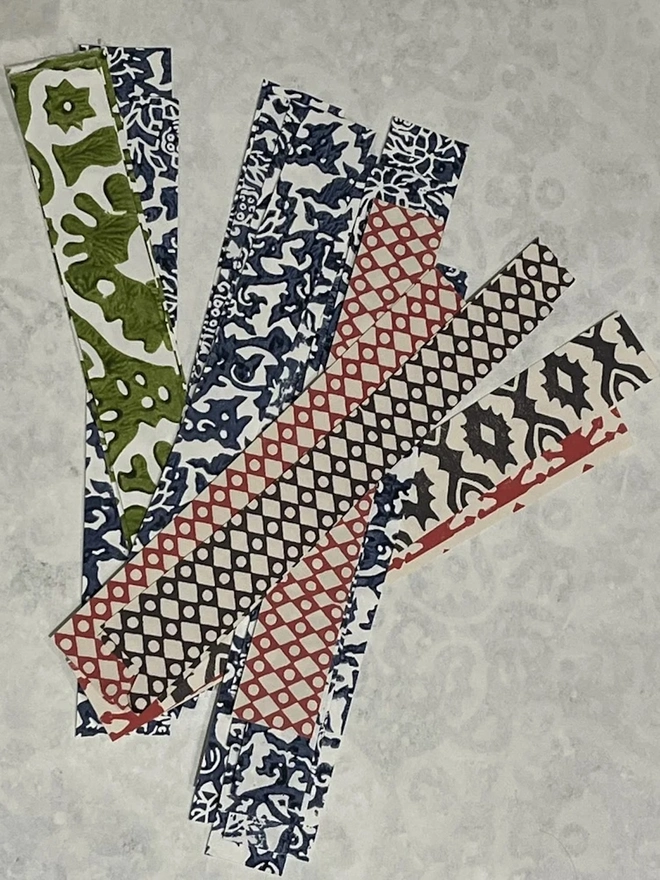 Colourful Paperchains (Set Of 60). Bundles of folded patterned paper strips in pink, green, navy and red, each held together with a contrasting paper band. These ready-cut strips are the pieces used to make a decorative paper chain garland.