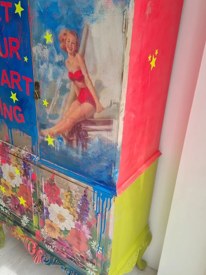 Upcycled cocktail cabinet in neons and bright colours. The decoration features a retro pin-up model on a boat and florals with stars and the wording reads ‘let your heart sing’