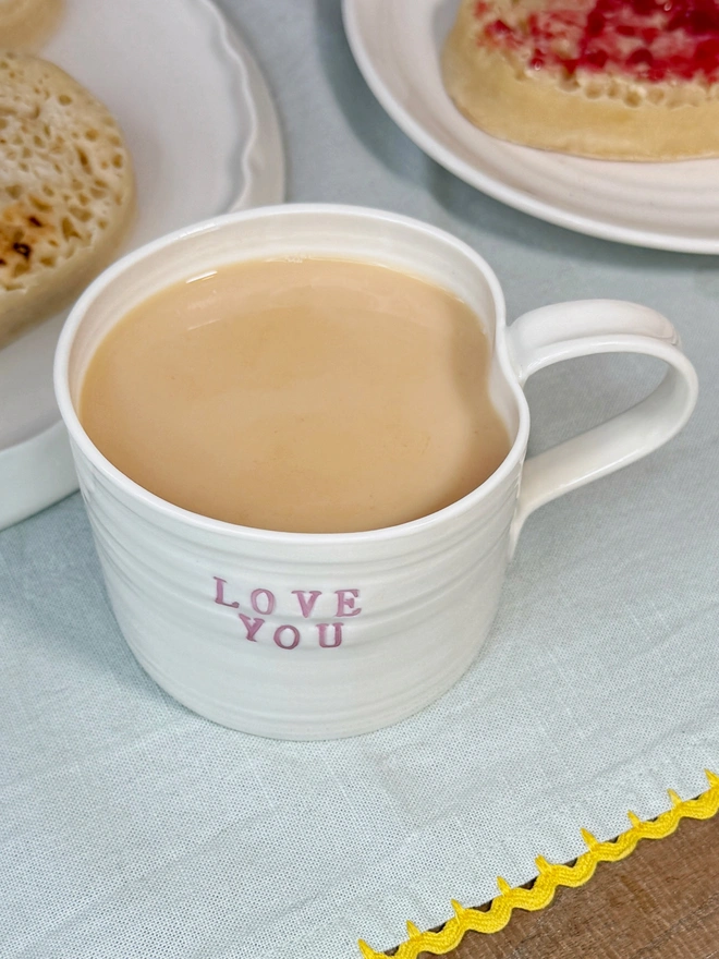 hand thrown porcelain cream mug with pink lustre text on the side that says LOVE YOU. The mug is sat upon a sage table cloth amongst a tea time scene of heart shaped crumpets