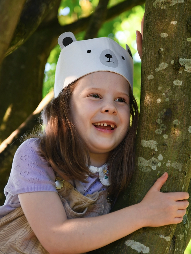 A young girl wearing a dress-up polar bear fabric crown and beige dungarees is climbing a tree.