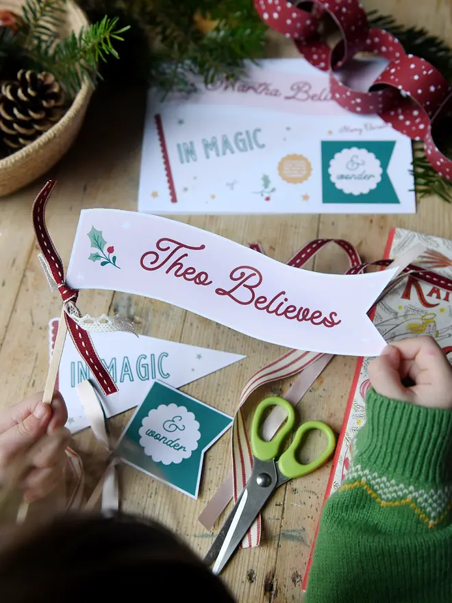 A young child is crafting Christmas flags with messages of magic and wonder printed on them. They are laid on a wooden desk with various traditional Christmas items around them.