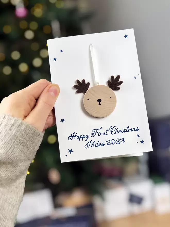A first Christmas greetings card with a small wooden reindeer decoration attached is being held in front of a Christmas tree.
