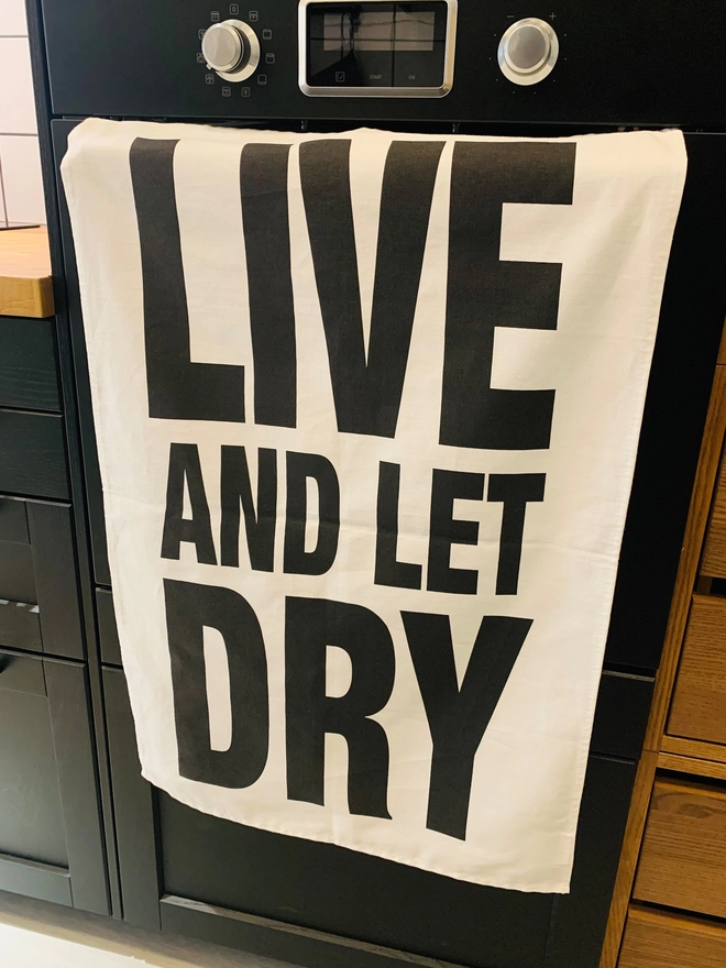 London Drying Live and Let Dry black text on white tea towel hanging on an oven door