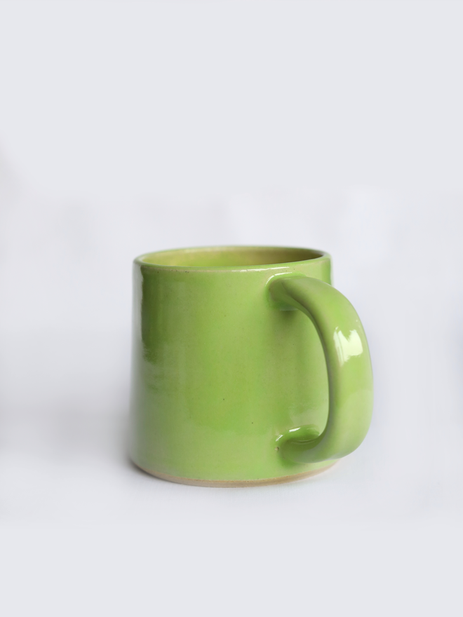 A handmade ceramic standard mug in glossy spring green glaze with a chunky loop handle, photographed against a plain light grey background