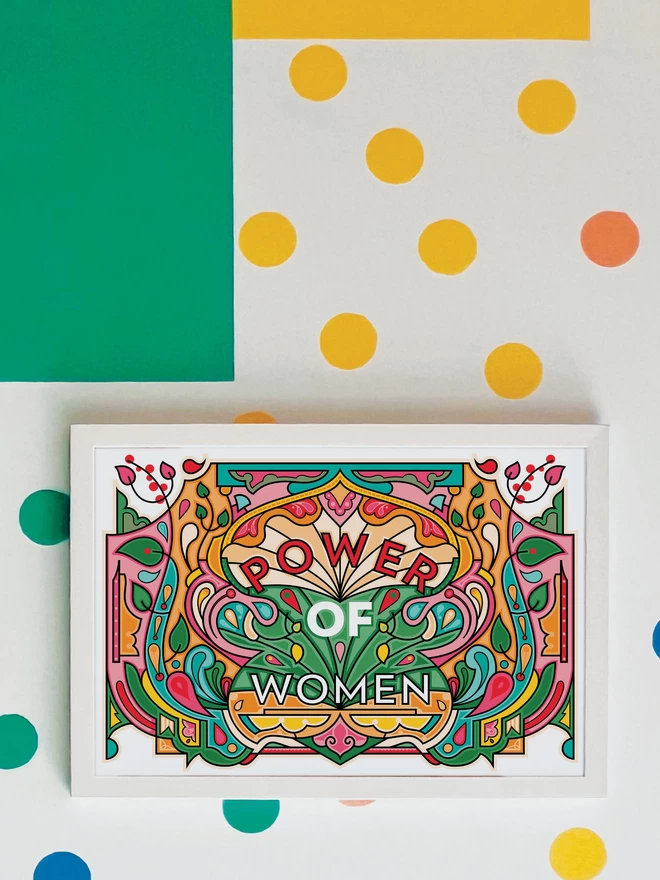 A vibrant, landscape illustration in greens, oranges and reds, with Power of Women written in the centre. The picture is hanging in a white frame on a white wall, with yellow, orange, green and blue spots and a green and yellow rectangle painted in the top left hand corner.