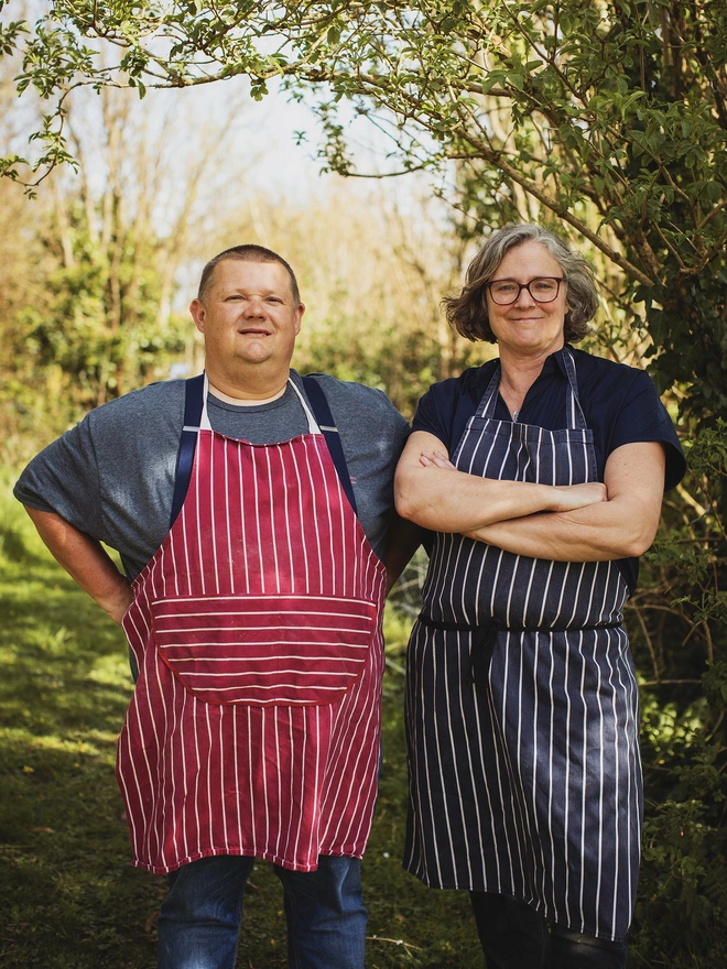 Man and woman wearing chefs aprons standing in a wood, smiling at the camera 