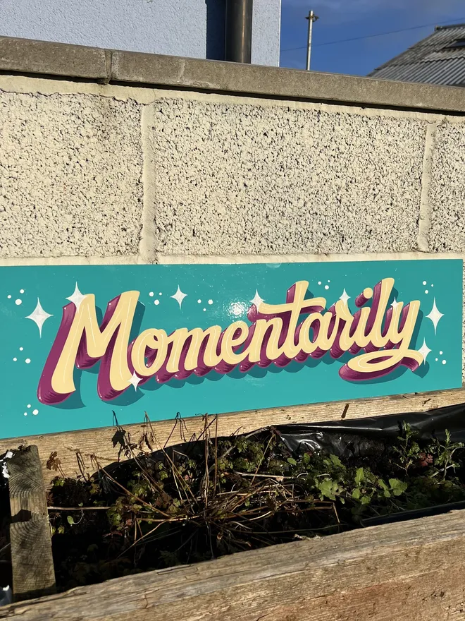 Turquoise background, ivory lettering and purple block shade lettering spelling 'Momentarily' on a hand-painted sign with white sparkle details..