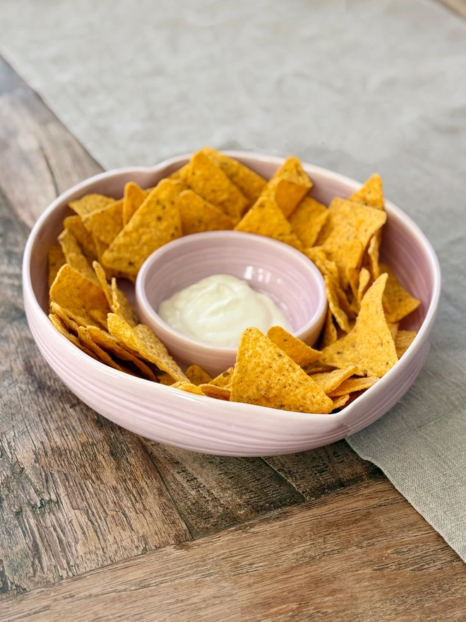 Heart shaped porcelain chip and dip bowl in pink glaze filled with tortilla chips and dip on a rustic wooden table