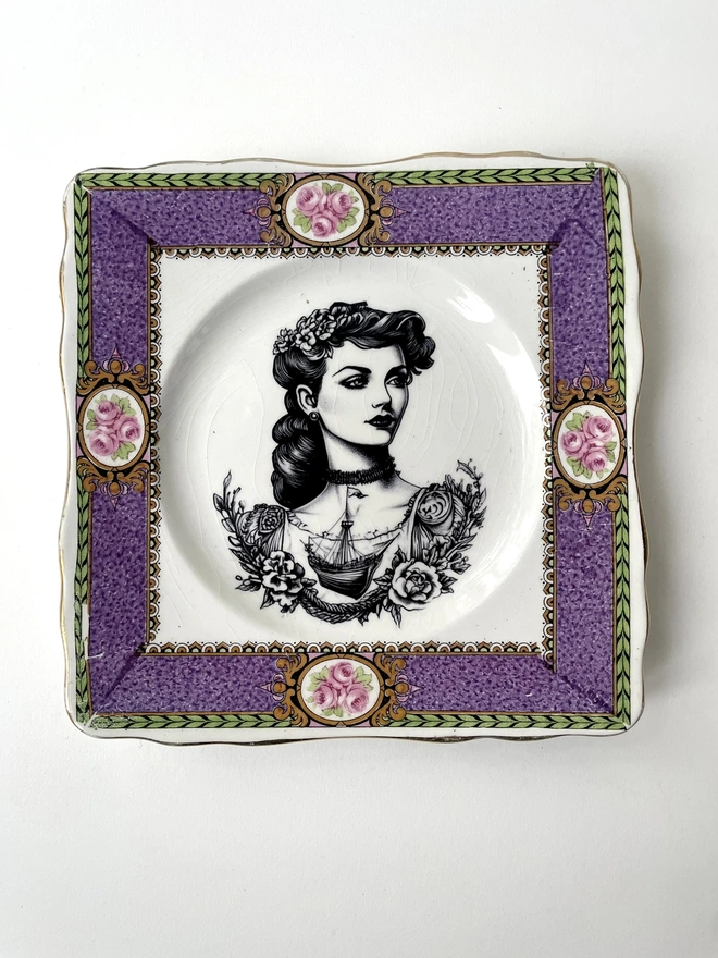 a beautifully ornate plate and in the centre is a vintage image of a beautiful tattooed london lady, she has ringlets and a choker and a tattooed ship on her chest