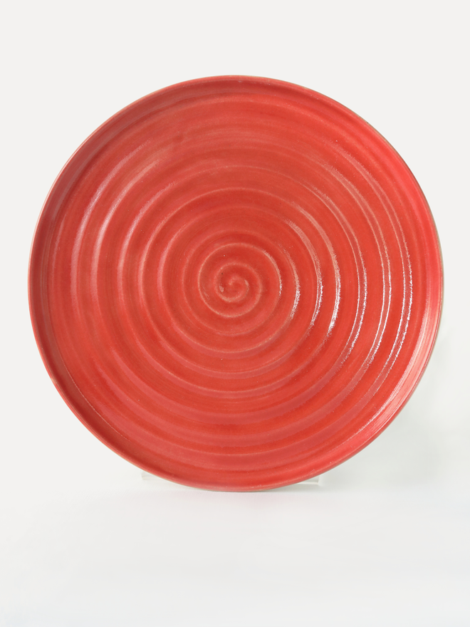 A handmade ceramic lunch plate in glossy cherry red glaze, photographed from above against a plain light grey background, showing a natural concentric swirl texture from the wheel-throwing process