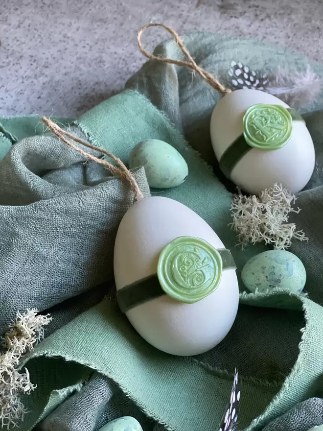 Twine string attacched to goose egg.  Green wax seal attached to green velvet ribbon around egg, Placed on soft ribbons and moss