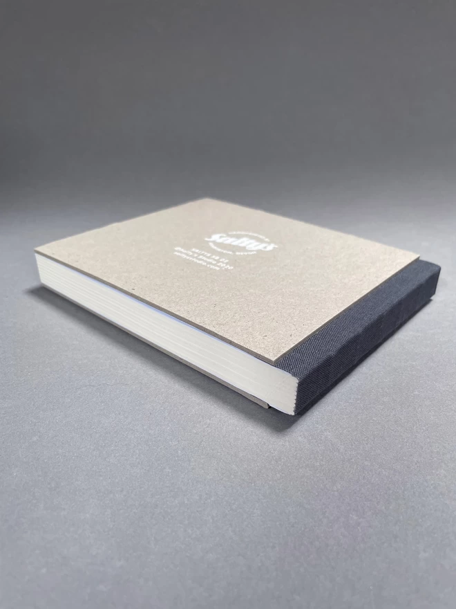 Reverse shot of a pasteboard sketchbook laid in a studio shot. Dark grey. Fabric spine and Saltys brand printed on reverse.