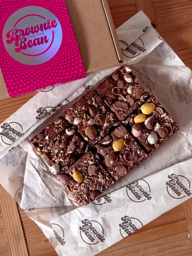 Easter Brookies (brownie top with a cookie base) on branded greaseproof paper with the branded box in the corner, atop a wooden worktop