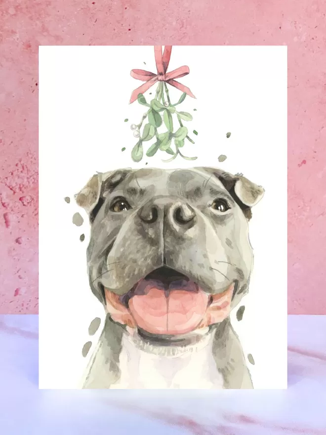 A Christmas card featuring a hand painted design of a Staffy, stood upright on a marble surface.