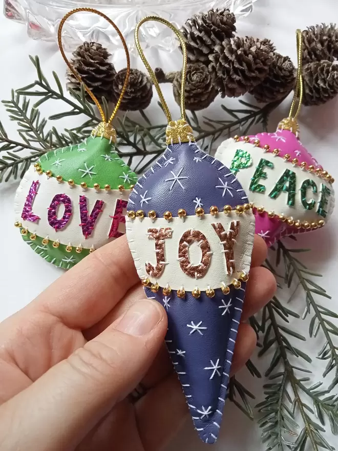 Three Hetty and Dave hand stitched retro inspired baubles hanging on a branch of fir, one is green with metallic pink lettering on a white band, spelling LOVE, one is purple with gold lettering spelling JOY, and one is pink with green metallic lettering spelling PEACE