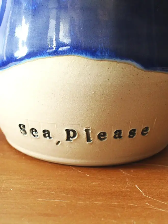 A 'Sea, please' ceramic mug with a blue glazed top and unglazed ceramic bottom resting on a wooden surface.