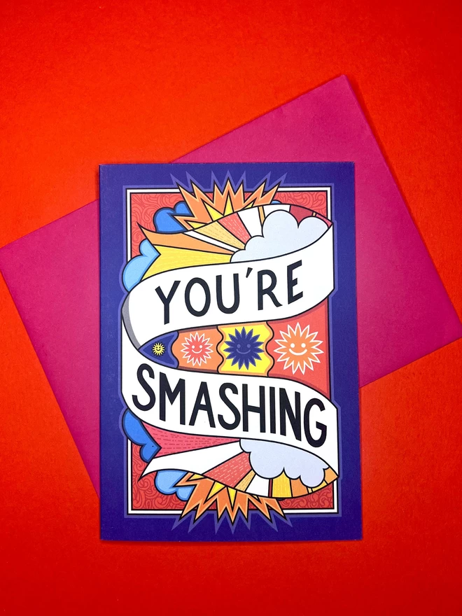A blue greetings card featuring “You’re Smashing” written in black on a white swirl across a vibrant design of the sky featuring clouds and drawings of the sun with smiling faces, sits on top of a pink envelope on a red backdrop.