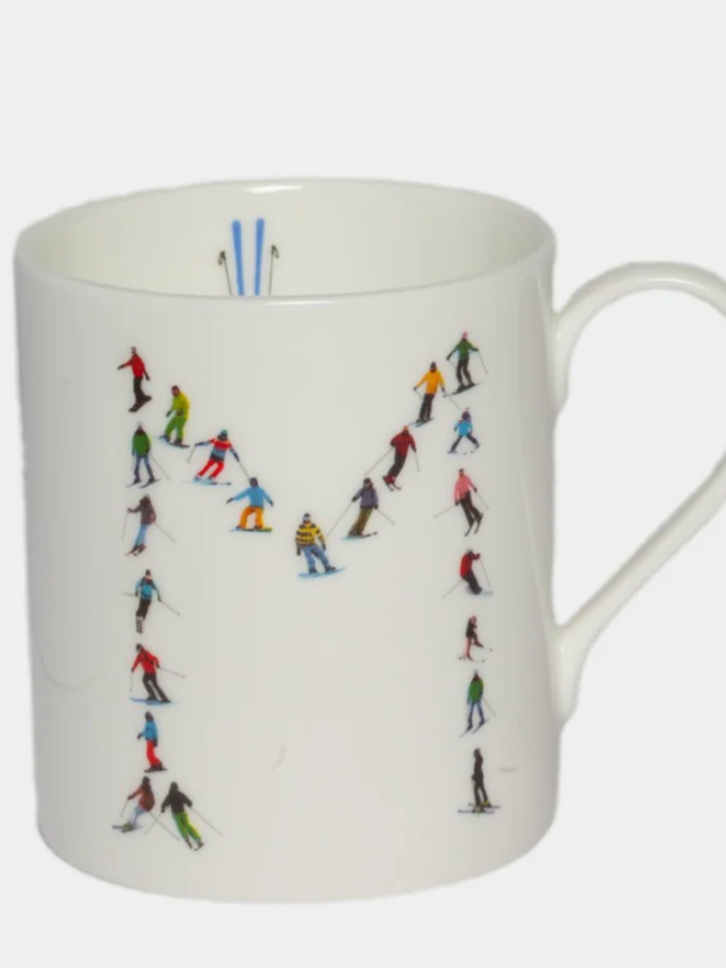A simple white mug illustrated with an ‘M’ made of small, multicoloured skiers. The mug is set against a plain, white background.