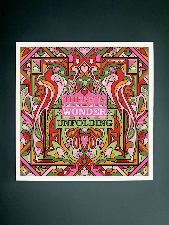 A vibrant, symmetrical design of pinks, greens and reds with There is Wonder Unfolding at the centre. The picture is hanging in a white square frame on a dark grey wall.
