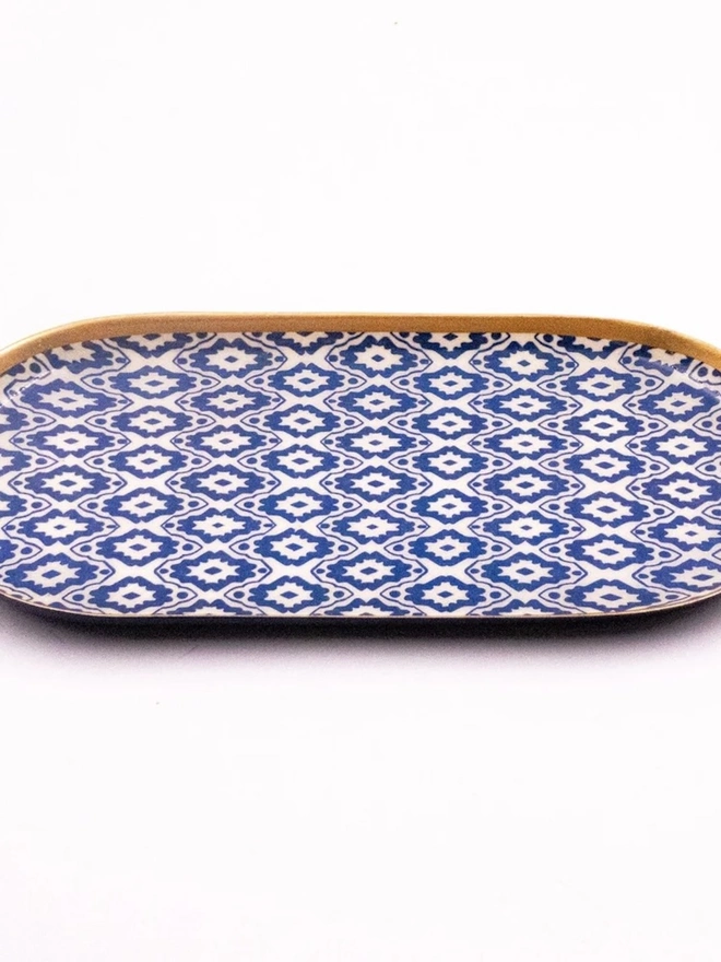 Perfect Enamel Oval Tray. An oval tray covered in blue and cream geometric ikat-style print with a polished gold rim, shown on a white background. The glossy patterned tray makes a stylish organiser for keys, jewellery or toiletries.
