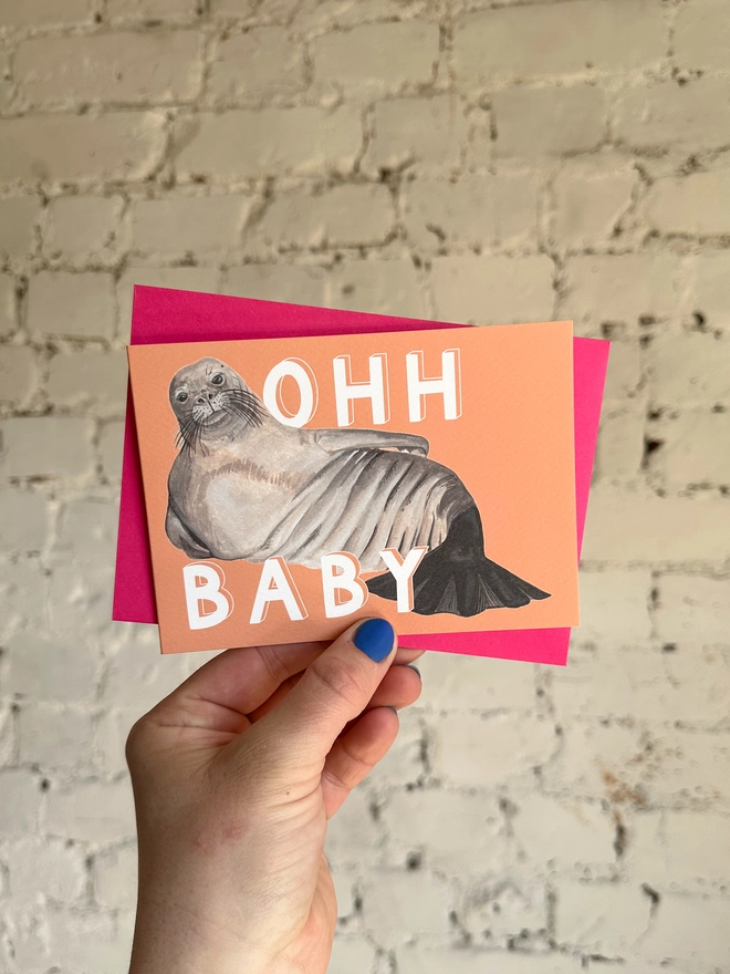 a peachy orange greetings card featuring an illustration of a chunky seal  with the text “ohh baby” with a neon pink envelope