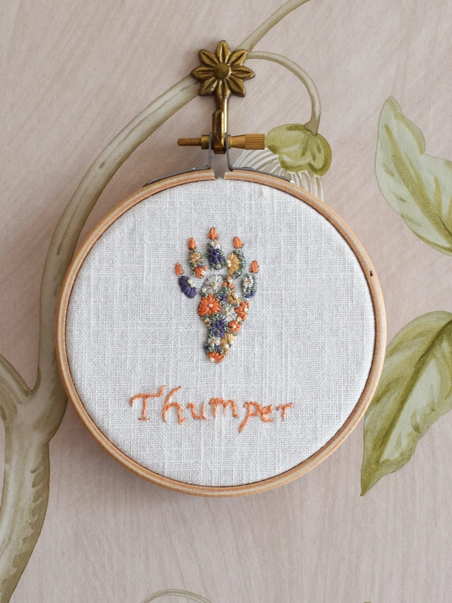 An embroidered Rabbits’ Foot of Golden Yellows and Bright Orange Blossoms with Green French Knot grass background.  Displayed in a hoop frame on a brass floral wall hook.