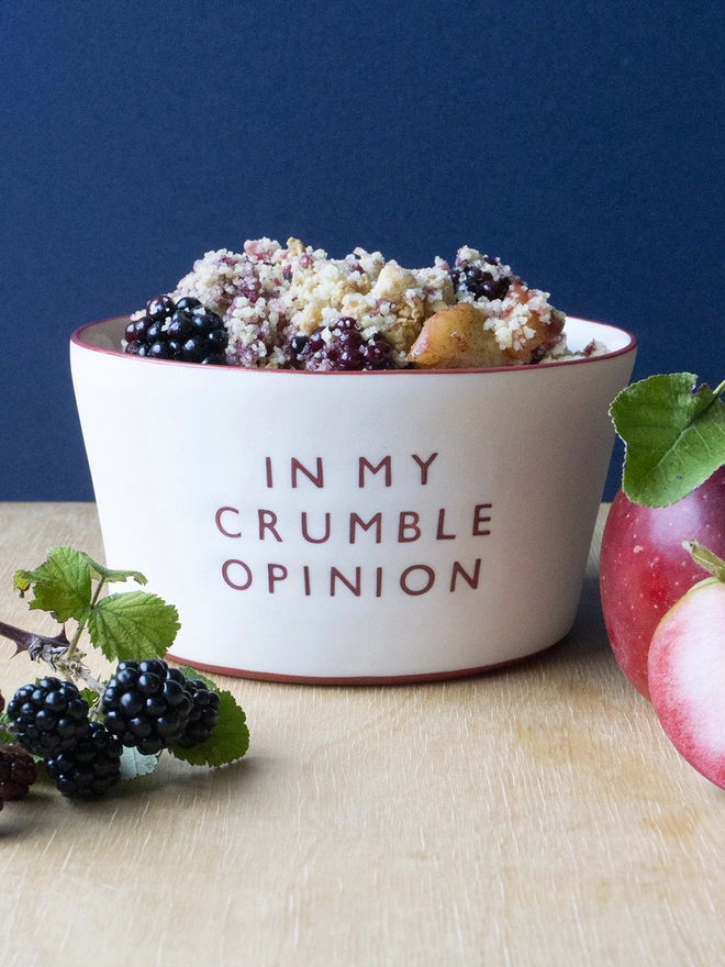 Ceramic porcelain pudding bowl with In My Crumble Opinion written on the side. A white pudding bowl with red rim containing apple and blackberry crumble next to blackberries and red apples.