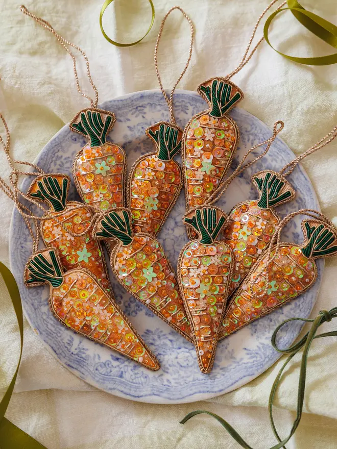 Multiple embroidered sequin carrot decorations laid on a pastel marbelled plate atop a white tablecloth, with green ribbon scattered around 
