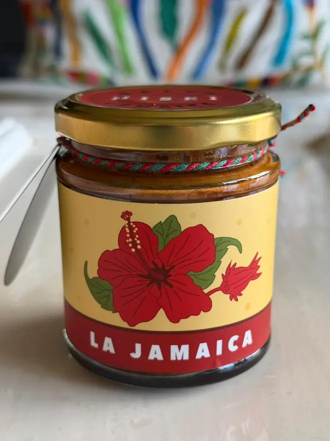'La Jamaica' Salsa Jar (Set Of 1-4 Jars), a jar of salsa sitting on a kitchen counter. It has the emblem of a flower on it. 