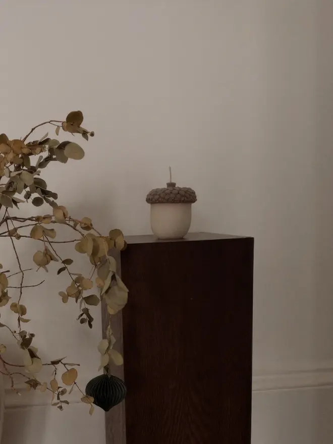 The Acorn Candle. An acorn-shaped candle with a textured taupe cap and cream base, displayed on a dark wooden plinth beside a branch of dried eucalyptus. The minimalist sculptural candle adds a calm, natural touch to autumn interiors.