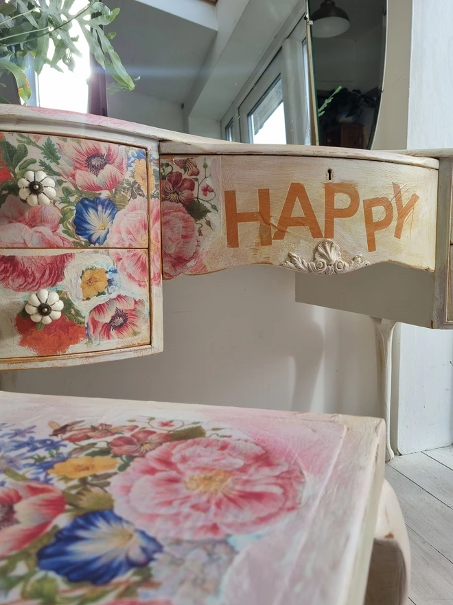 An antique kidney-shaped dressing table with chair and a trio of mirrors. Upcycled in cream and pale pink with decoupage florals in oranges and pinks. Text across the table read ‘flowers make me happy’ in the orange of the undercoat.