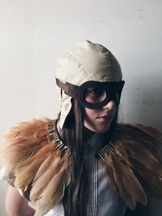 A child with long dark hair looking slightly to the left. they are wearing a beige cotton flying hat, brown leather flying goggles mask, and a copper coloured feather cape.  