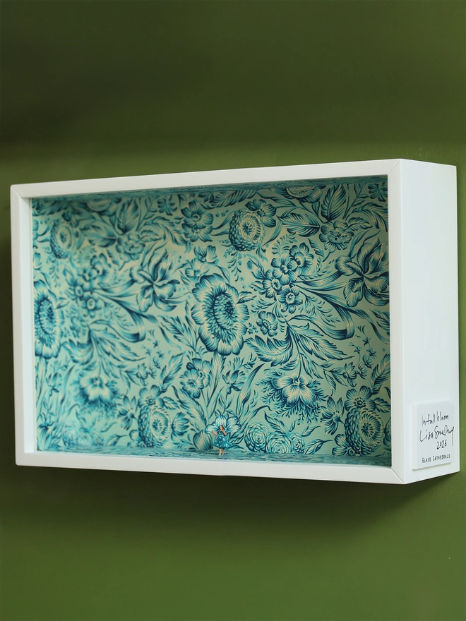 Miniature scene in an artbox showing a tiny lady in floral dress matching the wallpaper. Full box shown hanging on an olive green wall.