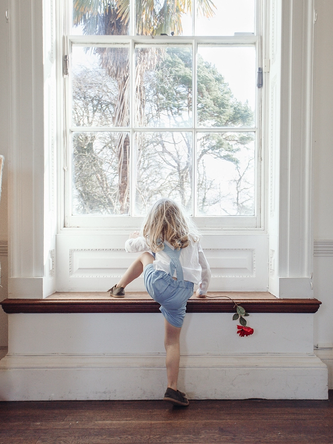 A boy in a white shirt and blue shorts with braces climbs onto a window seat