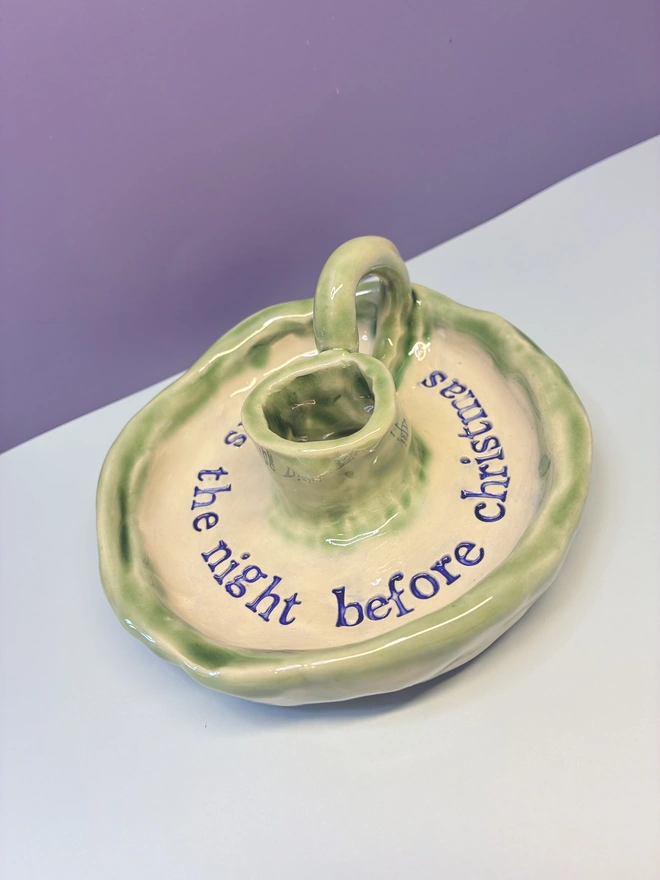 Handmade ceramic Christmas taper candlestick holder with a soft green glazed edge and the words “‘Twas the night before Christmas” in dark blue lettering, arranged around the candle holder opening. Photographed against a pale background.