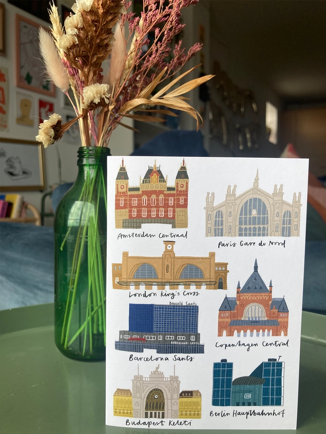 Greetings card with illustrations of UK train stations. Placed on a table with flowers in the background.