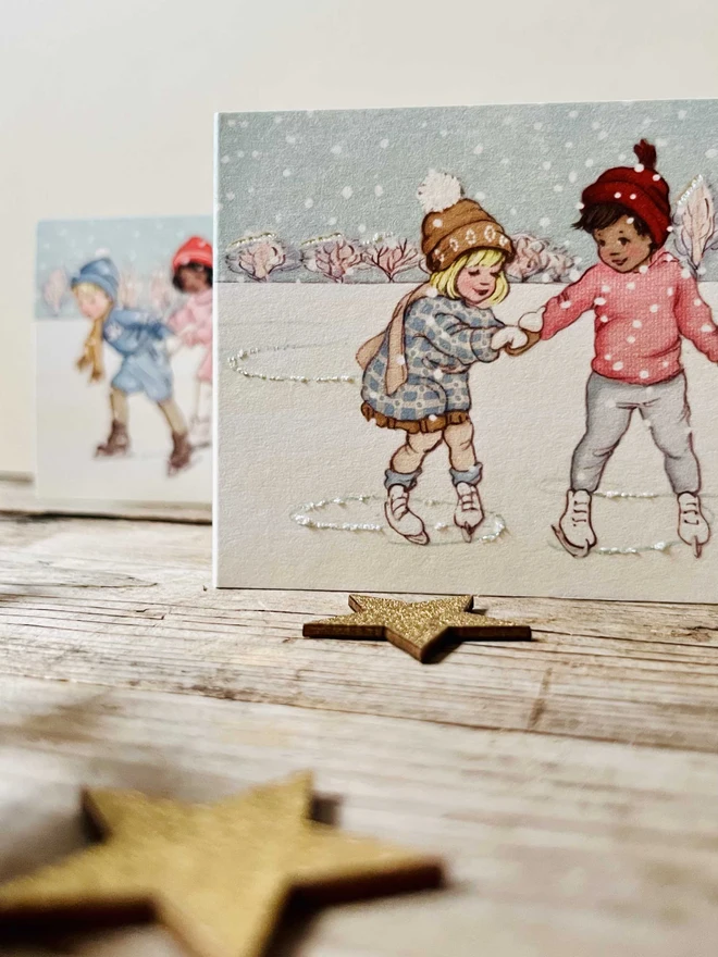 a close up of a retro style illustrated concertina cHristmas card featuring a little boy and girl learning to ice kate drawn in a retro vintage story book style hand finished with glitter