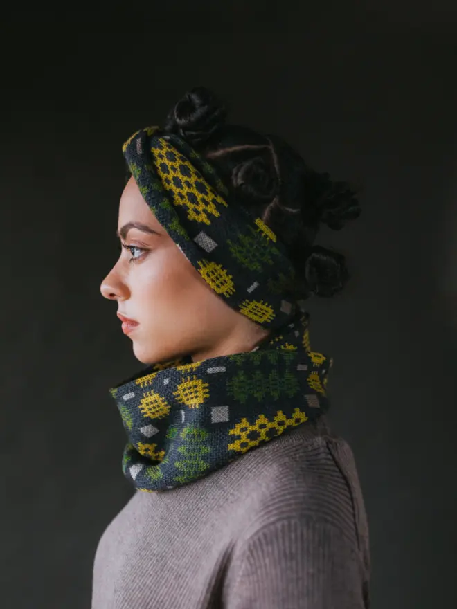 Model wearing pieces from MABLI's 'Carthen' collection, featuring the Carthen Wrap in the vibrant 'Anthracite/Pea' colour-way. The design showcases bold, colourful patterns inspired by traditional Welsh blanket tapestry.