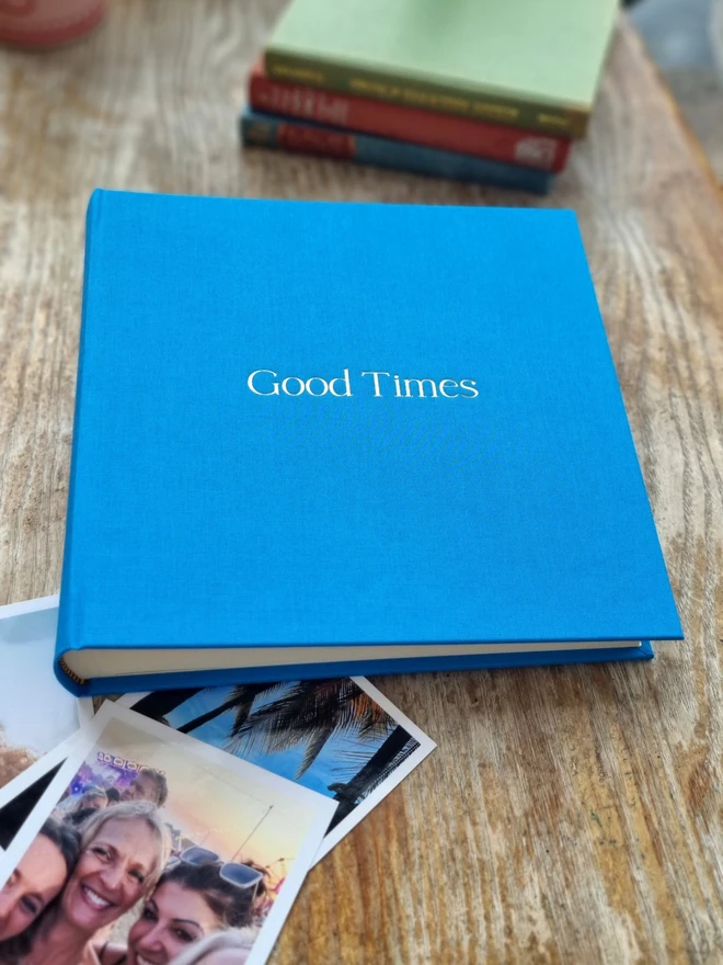 A sky blue photo album printed with the words 'Good Times' rests on a wooden floor and a rug, next to family photos.