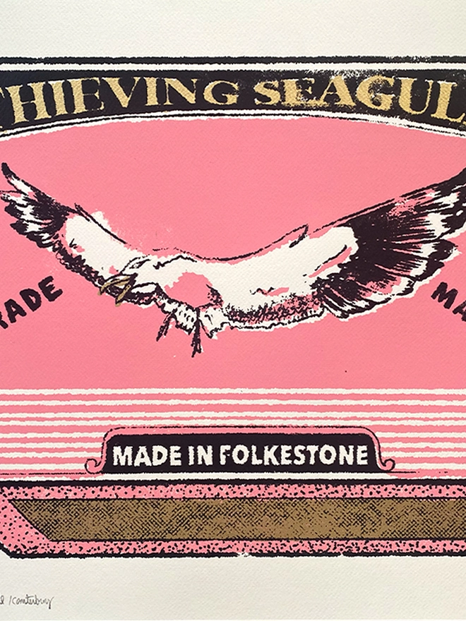 
A close-up photograph of a limited edition A2 screenprint. The print depicts a retro-style matchbox illustration in pink, black, and gold. A large, realistic seagull with a mischievous expression and an open beak is flying across the center. The words "THIEVING SEAGULL" are at the top, and "TRADE MARK" is on either side of the seagull. A banner at the bottom says "MADE IN FOLKESTONE." The background is a solid pink with a slight gold texture. The matches on the side are green and heart-shaped. Below the main image, the print is signed and numbered 3/13. The entire image has a grainy texture, like a vintage screen print.