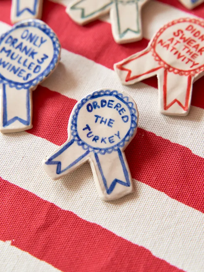 A selection of Red, Green & Blue Handmade Ceramic Christmas Award Badges, handpainted with different phrases, atop a striped red and white tablecloth