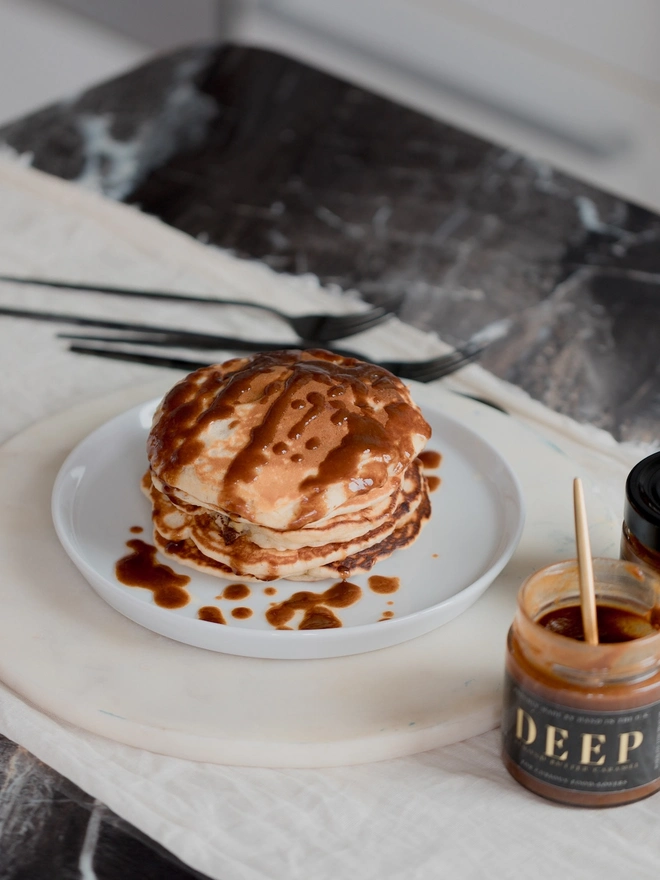 A jar of almond butter caramel sits in front of a plate of pancakes on a dark marbled surface with a white table runner