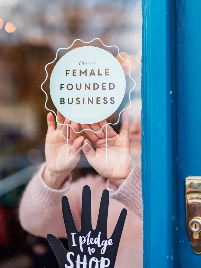 Female Founded Business Rosette Sticker on a shop door with a blue frame seen being put up by someone wearing a pink jumper.