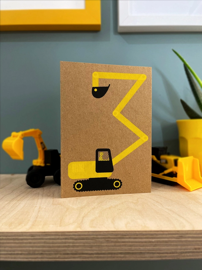 A brown kraft greetings card with a yellow and black digger stood on a plywood shelf, with a mini digger toy peeping from behind the card, and other framed pictures showing around the edges. A plant is half in shot.