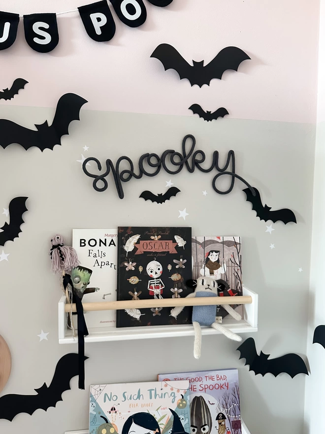 A 'spooky' knitted wire word wall sign hanging in a kid's playroom or bedroom above a reading area filled with Halloween books and cute Hallow's eve decor.