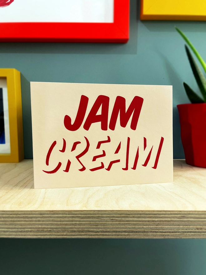 Cream at the bottom, Jam on top, printed in red ink on this cream card. Stood on a plywood shelf with edges of coloured frames showing around. 