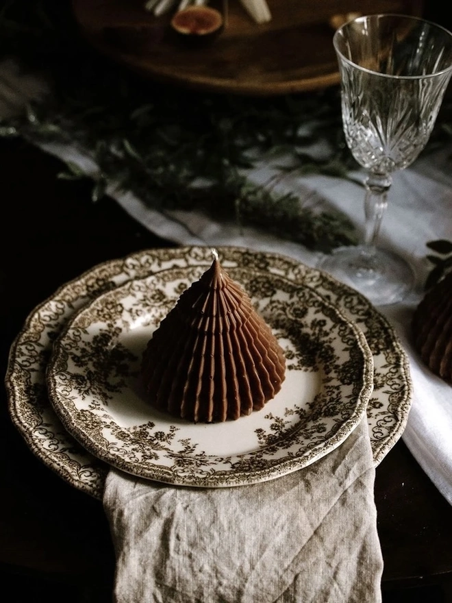 Brown Origami Christmas Tree Warm Candles (Set Of 2). A brown ribbed cone candle on a stack of vintage floral plates over a linen napkin, with a crystal glass and greenery on a dark table. The elegant candle doubles as a stylish Christmas place setting or table favour.