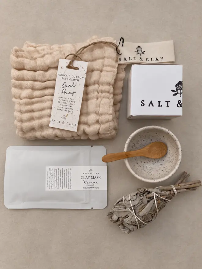 a cotton cloth with a sage smudge wand and a bowl and spoon in a flat lay image of the salt and clay calming rituals gift set.