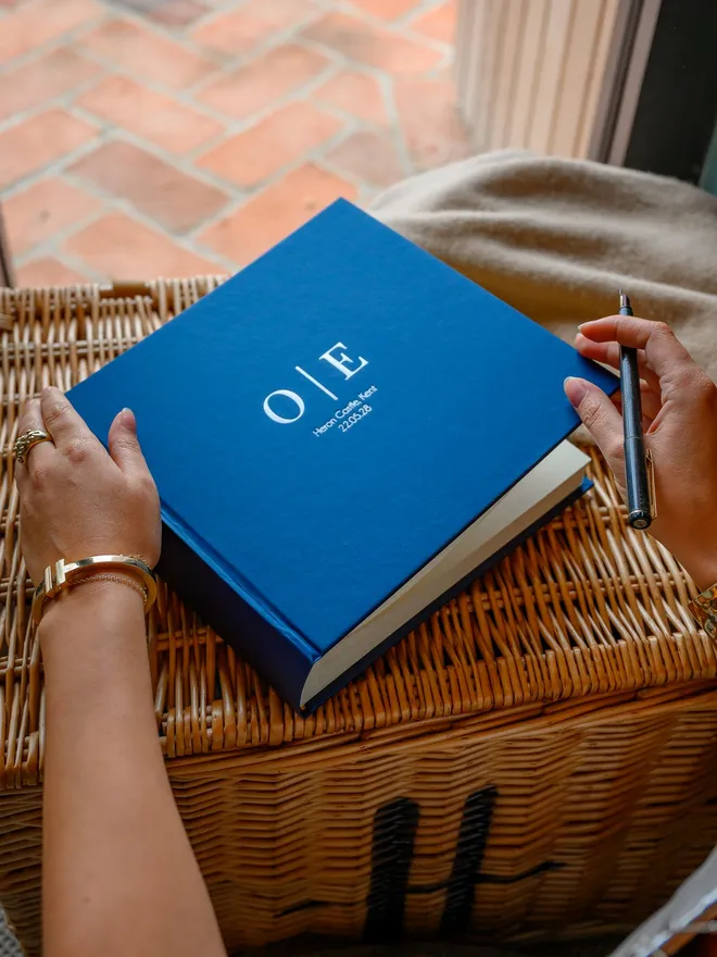 A blue hardback wedding guest book, printed with a couple's initials, rests on a wicker basket.